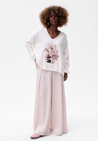 Vrouw met krullend haar, gekleed in een oversized witte trui met een bloemenprint, brede lichtroze broek, grote oorringen en roze schoenen, staand.