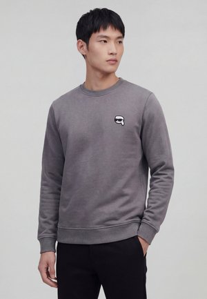 Young man wearing a gray sweatshirt with a small black-and-white cartoon face logo on the chest, standing with one hand in pocket.