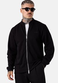 Schwarze Zip-Hoodie aus weichem Material, mit hohem Kragen, langen Ärmeln und silbernem Reißverschluss. Getragen über einem weißen T-Shirt.