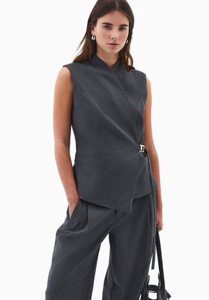 Ärmlös grå skräddarsydd väst med knyt-design, detaljer i silverfärgade spännen och sidofickor; matchad med lösa byxor.