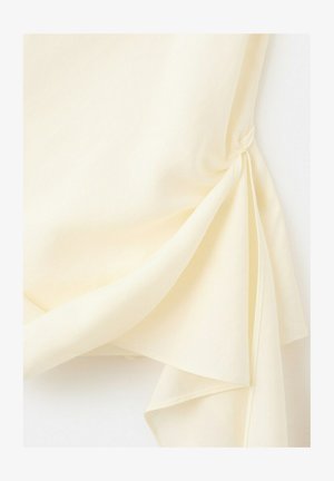 Krémová priehľadná látka prehodená a voľne zviazaná do uzla, vytvárajúc vrstvené záhyby a jemnú textúru na bielom pozadí.