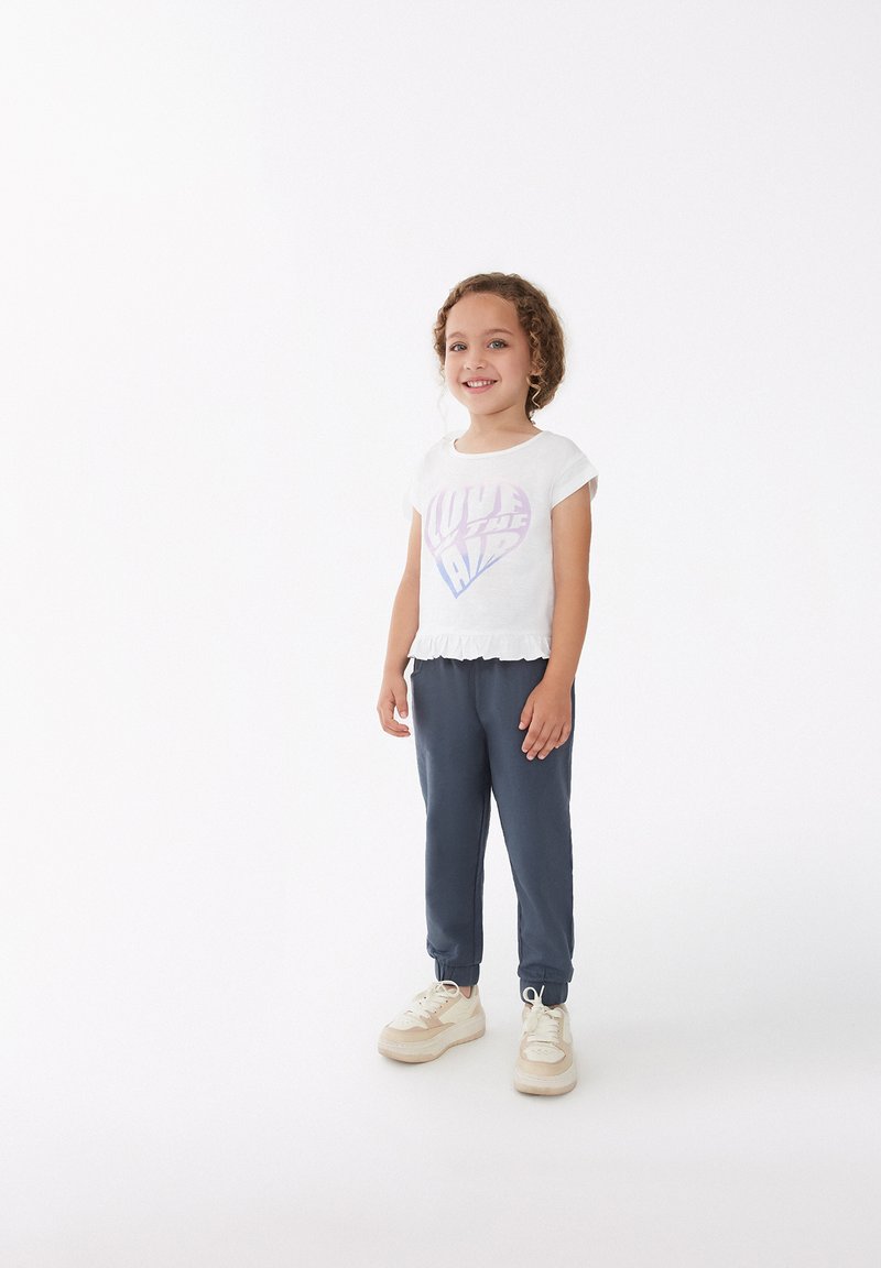 Camisa blanca de manga corta con un gráfico de corazón en tonos pastel y un dobladillo con volantes, combinada con pantalones navy ajustados y zapatillas beige con acentos blancos.