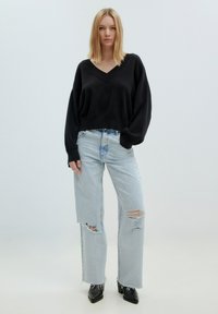 Schwarzer, übergroßer Pullover mit V-Ausschnitt und langen Ärmeln, kombiniert mit hellblauen, zerschlissenen, weiten Jeans. Schwarze, spitze Stiefeletten.