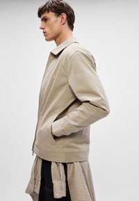 Beżowa kurtka zapinana na zamek, z kołnierzem, dwoma bocznymi kieszeniami i haftowanym logo. Posiada gładką teksturę oraz luźny krój.