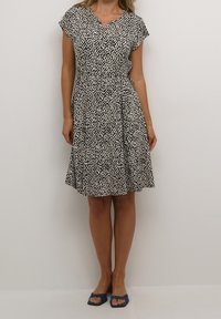 Mustavalkoinen kuvioitu mekko, jossa on hihat, V-pääntie, vyötäröstä kiristetty ja liehukeleikkaus. Päälle on puettu siniset avonaiset sandaalit.