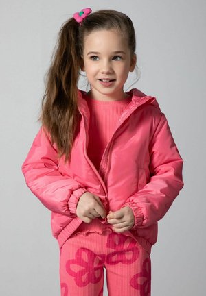 Dziewczyna o długich brązowych włosach związanych różową opaską w kwiaty, ubrana w jaskraworóżową kurtkę i pasujące spodnie w kwiatowy wzór, zapinająca suwak kurtki.