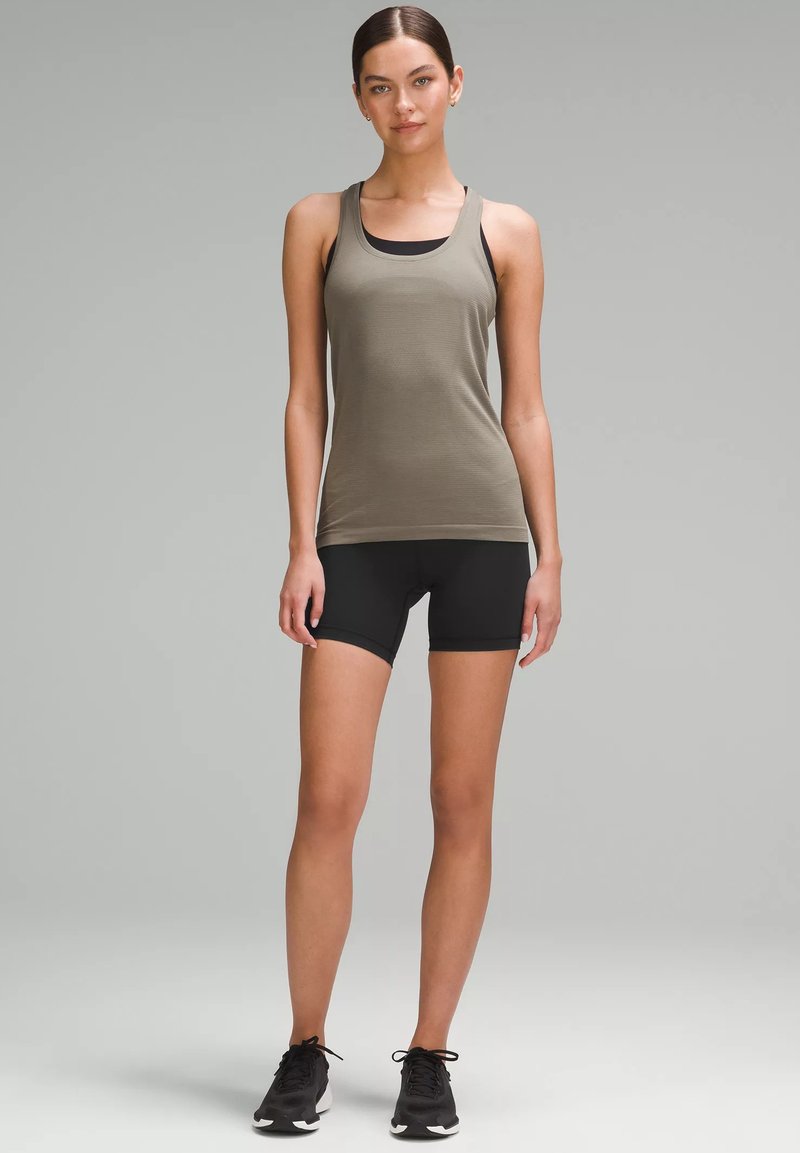 Top da allenamento verde oliva con scollatura rotonda, abbinato a pantaloni corti neri aderenti e scarpe da ginnastica nere. Tessuto liscio e leggero.