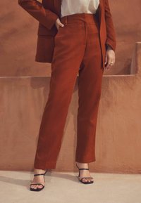 Pantalons en lin couleur rouille avec une coupe ajustée et des jambes droites, associés à des sandales noires à lanières. Texture lisse, design minimal.