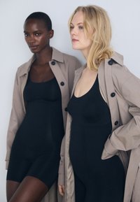 Two models wear fitted black bodysuits. They are layered with light beige trench coats, characterized by large buttons and a relaxed fit.