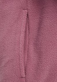 Fleecejack in een gedempte rozekleur, met een zachte textuur en een ingenaaide zijzak. Het materiaal oogt zacht en warm.