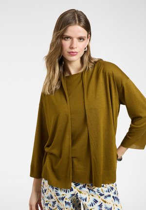 Jonge vrouw met steil lichtbruin haar, gekleed in een olijfgroene cardigan over een bijpassende top en een witte, blauwe en gouden broek met patroon.