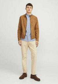 Ruda zomšinė striukė su užtrauktuku, derinama su mėlyna dryžuota marškinėliais, kremiškos spalvos kelnėmis ir rudais raištiniais batais. Paprastas dizainas, modernus kirpimas.