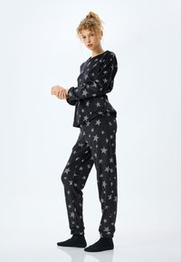 Musta pyjama-asuste, jossa on hopeisia tähtikuvioita, pitkät hihat ja kapeat housut, valmistettu pehmeästä kankaasta, jossa on sileä rakenne.