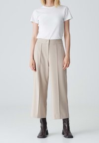 Pantaloni beige a gamba larga con pieghe frontali e chiusura a un bottone, abbinati a una t-shirt bianca aderente e stivaletti neri alla caviglia.