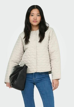 Kvinde med langt mørkt hår iført en lys quiltet jakke og blå jeans, der holder en stor sort håndtaske ved siden af sig.