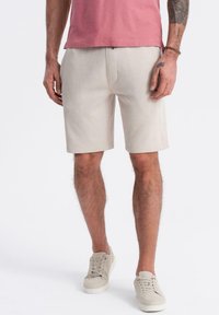 Shorts beige clair en tissu doux, avec une coupe droite, des poches latérales et une ceinture. Portés avec des baskets de couleur claire.