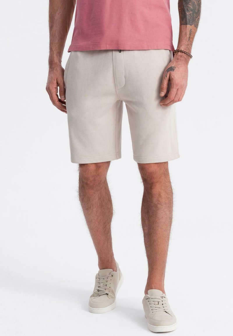 Shorts beige clair en tissu doux, avec une coupe droite, des poches latérales et une ceinture. Portés avec des baskets de couleur claire.