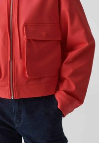 Rote Bomberjacke mit einem Frontreißverschluss und einer großen Brusttasche. Sie hat lange Ärmel und eine lockere Passform, ergänzt durch eine dunkle Jeans.