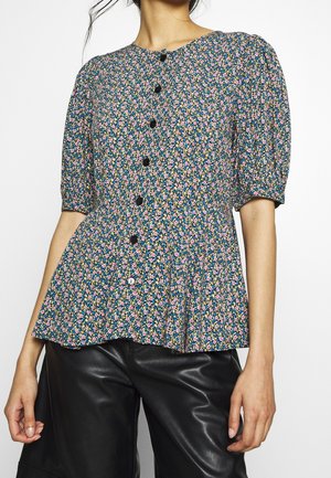 Kvinde iført en blå blomsterprintet bluse med knapper og pufærmer samt sorte læderbukser, vist fra skuldre til hofter.