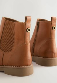 Une paire de bottes chelsea en cuir beige avec des panneaux élastiques sur les côtés, des languettes et des semelles en caoutchouc, présentée de l'arrière sur un fond clair.
