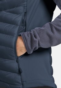 Marineblaue Jacke mit gestepptem Material auf einer Seite und glattem Stoff auf der anderen Seite, versehen mit einer Reißverschlusstasche und einem gerippten Ärmelbund.