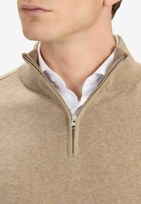 Hellbeiger Zip-Up-Pullover mit Stehkragen, gerippter Textur und metallischem Reißverschluss, übereinander getragen mit einem weißen Hemd.