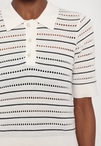 Knit polo shirt in cream with black horizontal stripes. Features a collar and three pearl buttons. Textured with open stitch patterns.