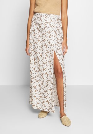 Donna con top beige senza maniche e gonna lunga bianca a stampa floreale con uno spacco alto sulla coscia, abbinata a scarpe beige intrecciate slip-on.