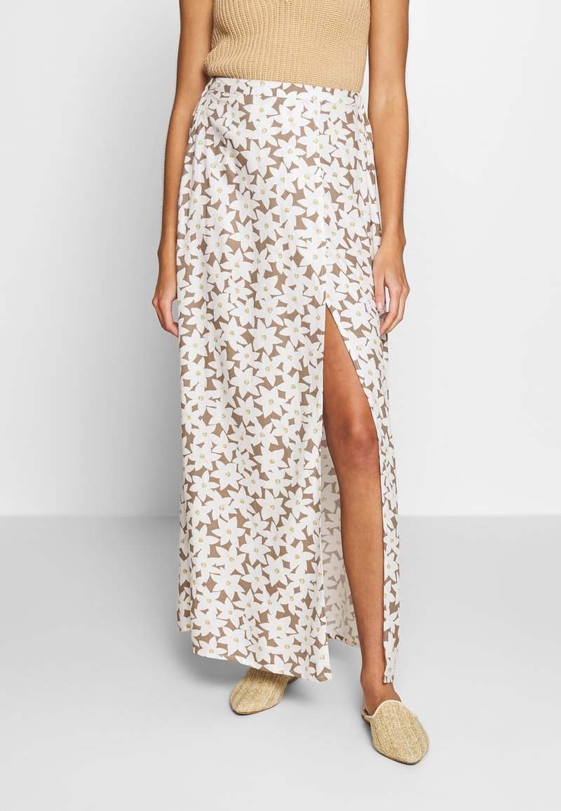 Femme portant un haut beige sans manches et une jupe longue blanche à imprimé floral avec une fente haute à la cuisse, associée à des chaussures beige tissées et sans lacets.