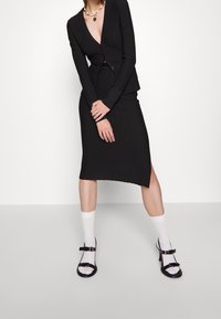 Svart ribbad figursydd kofta med V-ringning, kombinerad med en matchande kjol med sidospjälkning. Bärs med vita strumpor och svarta sandaler med remmar.