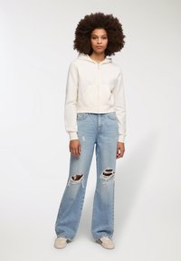 Bawełniana bluza z kapturem w kolorze off-white, z kieszonką z przodu, w połączeniu z jasnoniebieskimi szerokimi spodniami z przetarciami i kremowymi butami.