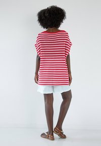 Czerwono-biały, pasiasty, oversize'owy sweter, luźny krój, połączony z jasnymi szortami. Brązowe sandały z detalami na paskach. Gładkie białe tło.