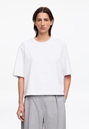 Person with medium-length dark hair wears a plain white short-sleeve top and light gray pleated trousers, standing against a white backdrop.