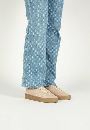 Baskets en suède beige avec des semelles en caoutchouc marron clair, portées avec un jean en denim bleu présentant un motif texturé de petites formes blanches.