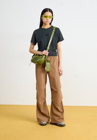 T-shirt noir à manches courtes, pantalon beige à jambes larges, sac bandoulière vert, lunettes de soleil vertes et chaussures à motif zèbre sur un tapis jaune.