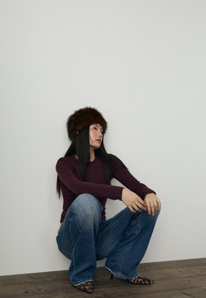 Rjava krznena kapa, dolgi rokavi bordo zgornji del, svetlobluje flare jeans in petke s leopardjim vzorcem. Lesena tla in enostavna bela stena v ozadju.