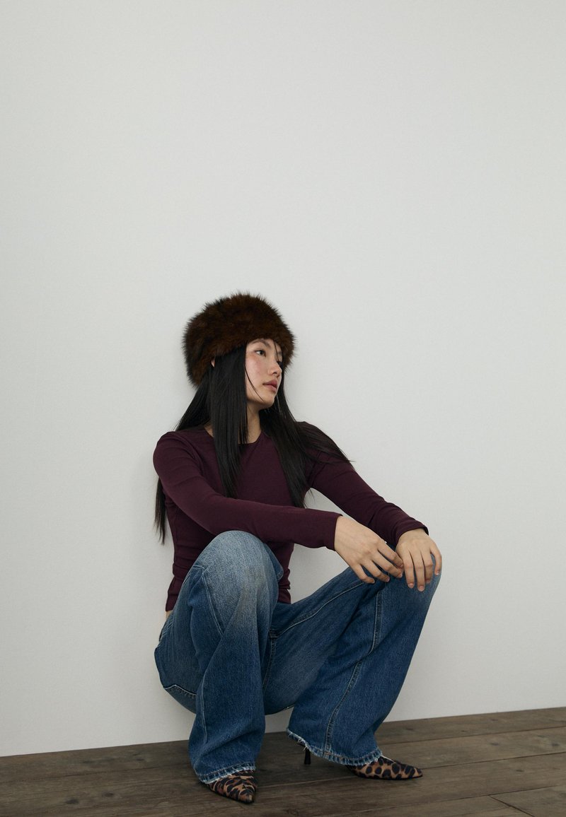 Chapeau en fourrure marron, top à manches longues bordeaux, jeans évasés bleu clair et talons imprimé léopard. Sol en bois et mur blanc uni en arrière-plan.