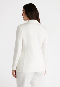 Biała marynarka wykonana z gładkiego materiału, o dopasowanym kroju, długich rękawach i minimalistycznym designie bez widocznych elementów metalowych.