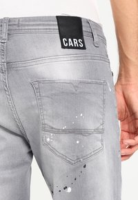 Grå denimjeans med en svart "CARS"-etikett. Har en distressed yta med färgstänk och standard bakfickor.