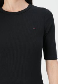Zwart geribbeld T-shirt met korte mouwen en een ronde halslijn, voorzien van een klein logo met rode, witte en blauwe accenten op de linkerkant van de borst.