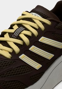 Pruun sportjalanõu võrgutäidisega, kollaste paelte ja valgete detailidega. Omaduseks on tekstuuriga disain ja silmapaistev bränding.