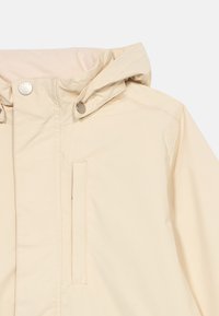 Μπεζ μπουφάν με κουκούλα, με κουμπιά σναπ στο γιακά και κάθετη τσέπη με φερμουάρ στο στήθος, κατασκευασμένο από λείο ύφασμα.