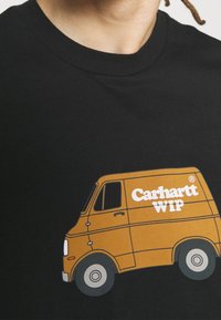 Persona vistiendo una camiseta negra con un gráfico de una furgoneta naranja y el texto blanco "Carhartt WIP" en el pecho, cortada en el cuello y los hombros.