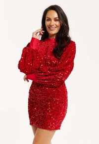 Red sequin dress with long, puffed sleeves and a high neckline. Fitted silhouette with a textured, shimmering surface.