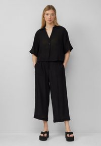Zwarte, loszittende, cropped blouse met kraag en knoopsluiting, gecombineerd met brede zwarte broek, beiden met een gekreukt oppervlak.