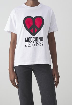 Modellen bærer en hvid T-shirt med et rødt hjerte og fredssymbol, hvor "MOSCHINO JEANS" er trykt nedenfor, parret med mørke jeans.