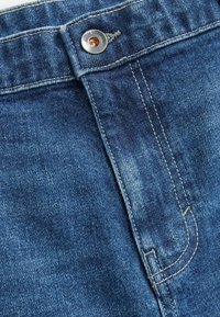 Sinised denim püksid, millel on metallist nööp sulgemine, nähtavad tikandid ja veidi pleekinud tekstuur. Suumvaade vöökohale.