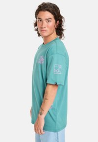 Hedvábně modré bavlněné tričko s krátkými rukávy, na přední straně a rukávu má růžové grafické designy. Standardní střih s kulatým výstřihem.