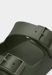 Ελαστικό σανδάλι σε ελιά πράσινο, με δύο ρυθμιζόμενες αγκράφες και ανοιχτή σχεδίαση στο δάχτυλο, που διαθέτει μινιμαλιστικό στιλ και ομαλή υφή.