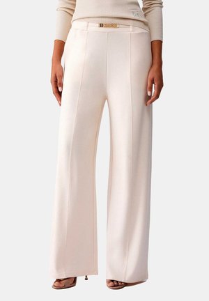 Pantaloni beige a vita alta a gamba larga con cuciture frontali, cintura decorativa in catena color oro, indossati con un maglione a coste abbinato e tacchi aperti.